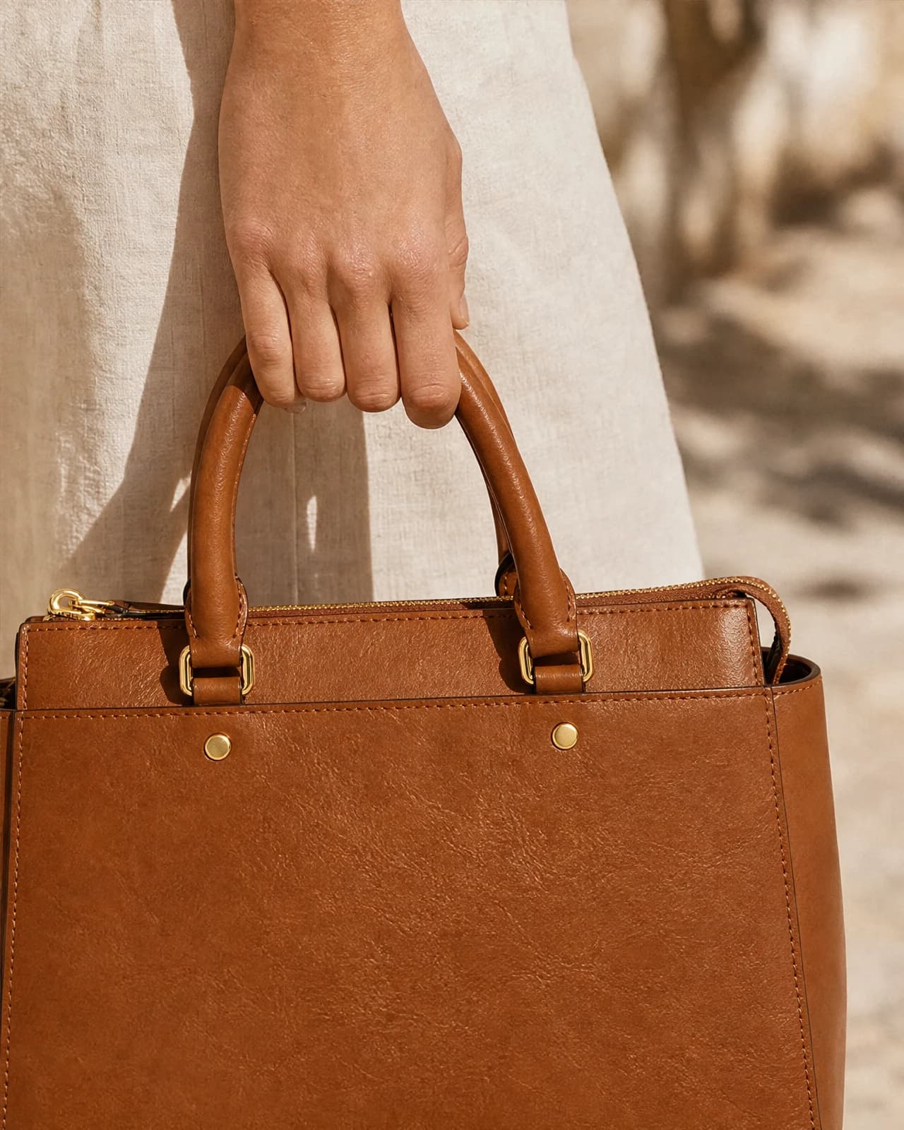 Macro detail of a hand lifting the tan leather handbag by its top handles.
