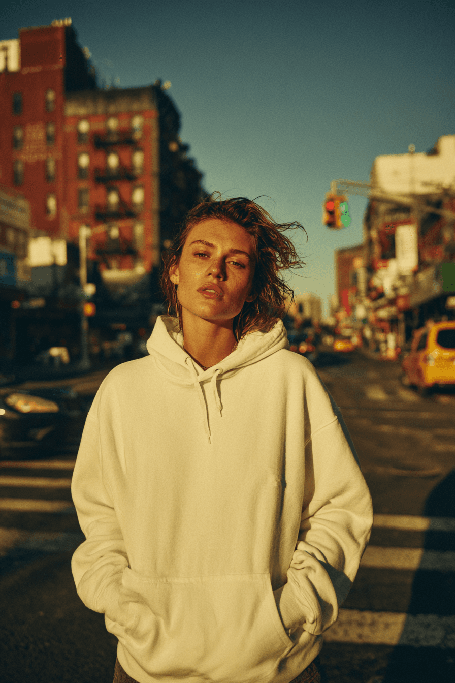 A slider that helps the user compare before and after images after being edited with nightjar. On the left side, there is a woman standing in a street and wearing a white hoodie. On the right side, there is the AI generated image which shows the exact same woman, standing in the same pose and in the same street, but the only difference is that now she is seamlessly wearing a leather jacket that looks 100% real and natural. - Before transformation