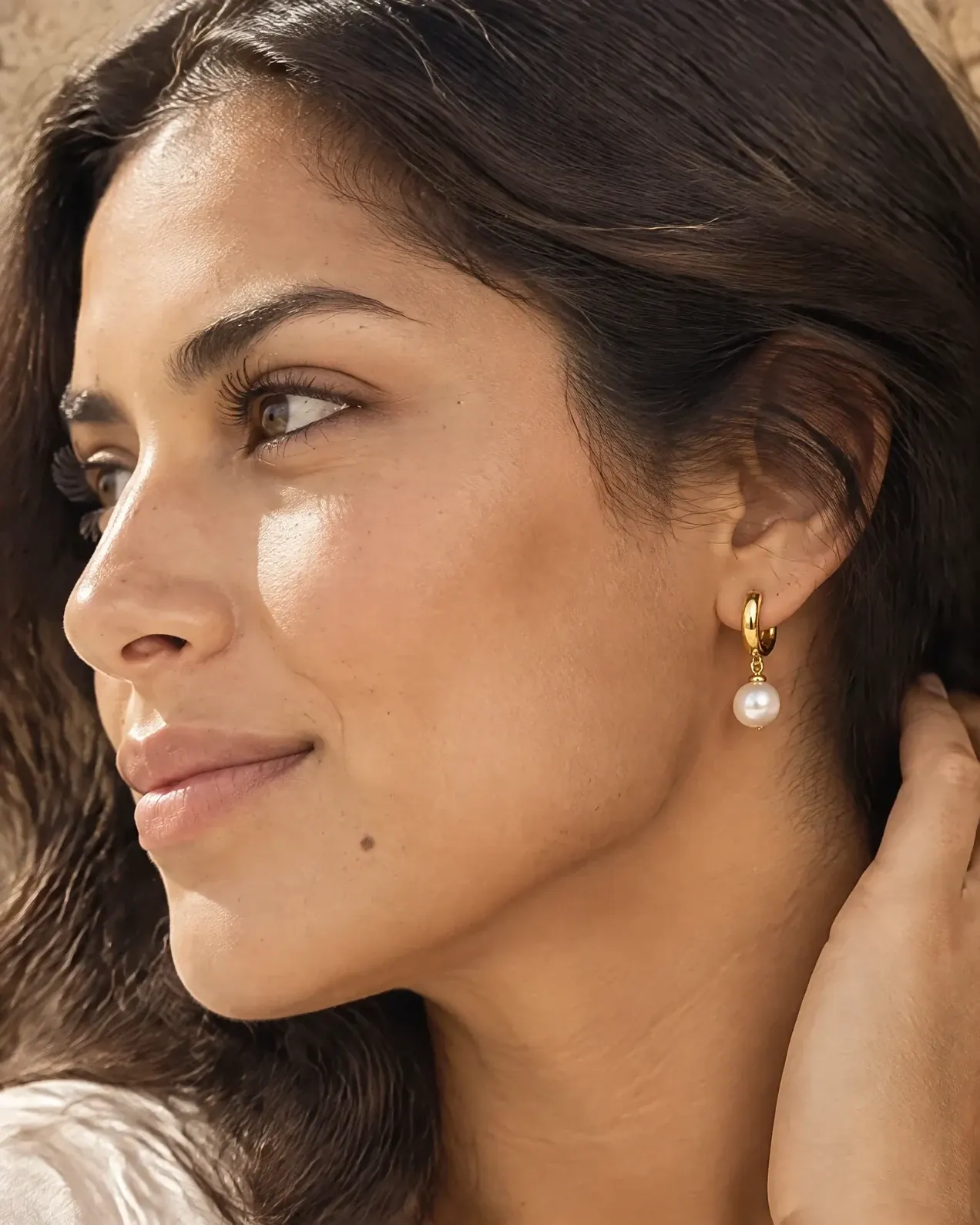 A model in profile wears a small gold huggie hoop earring with a pearl drop in soft daylight.