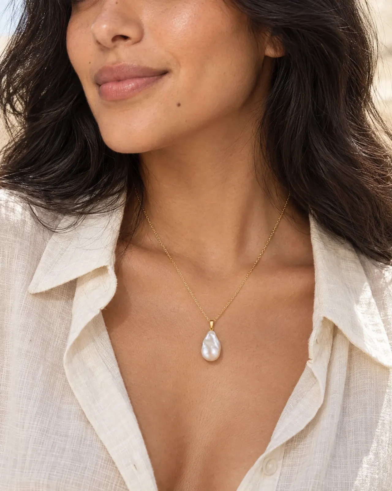 A close crop of a model in an open ivory linen shirt wearing a baroque pearl pendant on a fine gold chain.