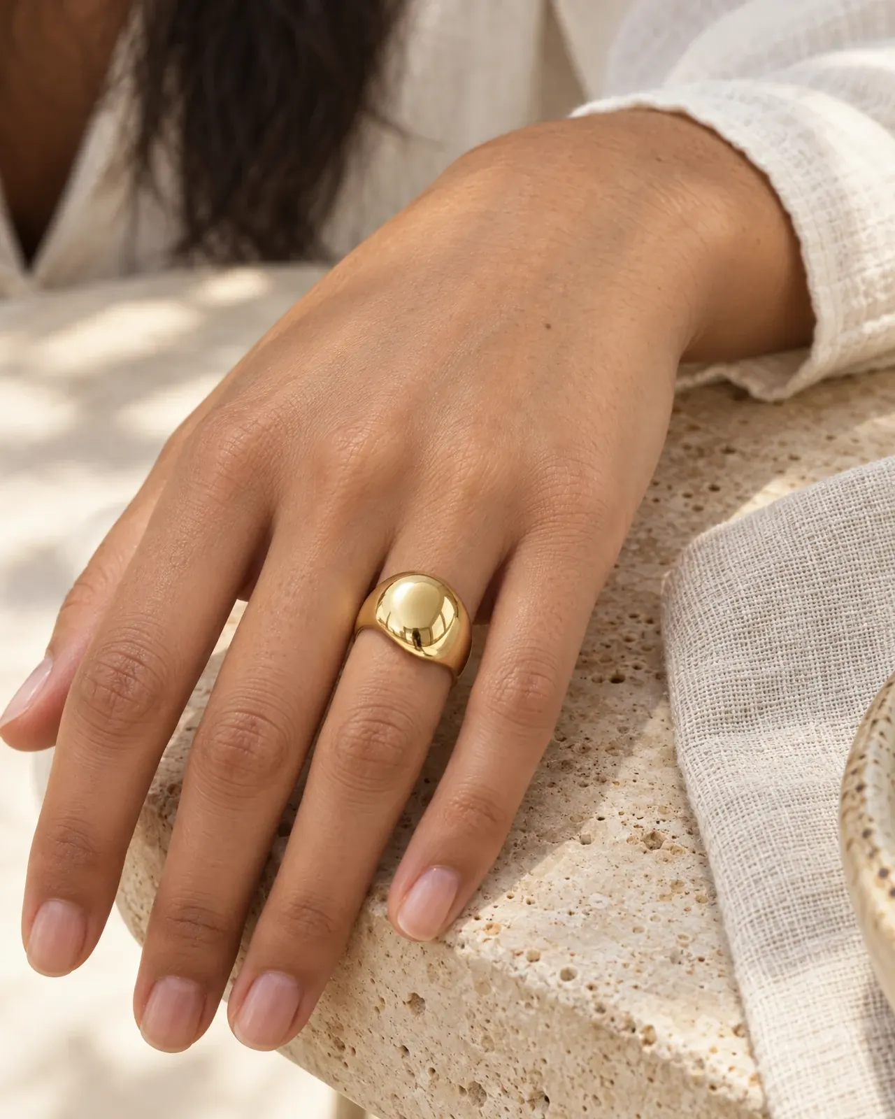 A hand resting on a travertine table beside linen, wearing a polished gold dome ring.