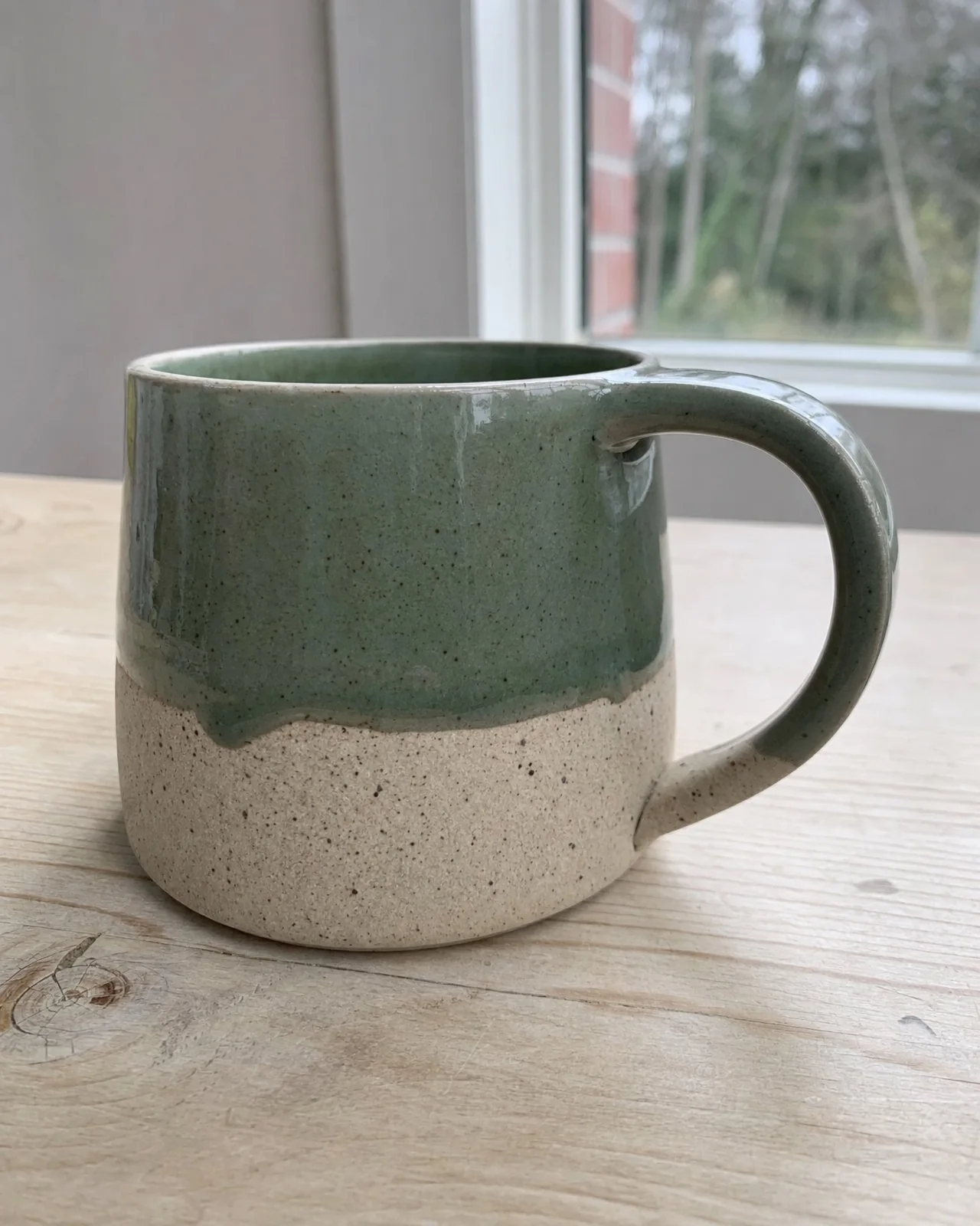 A phone photo of a speckled stoneware mug with a sage green glaze on a wooden table.