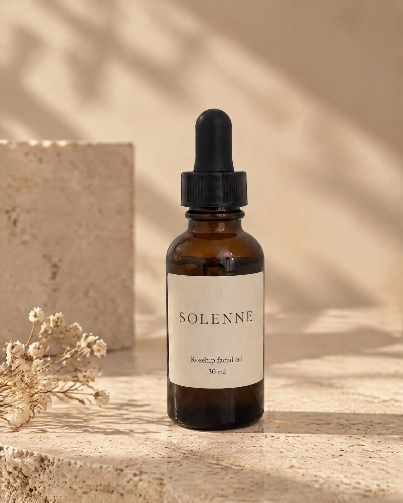 The rosehip oil bottle on a travertine ledge beside dried flowers in morning light.