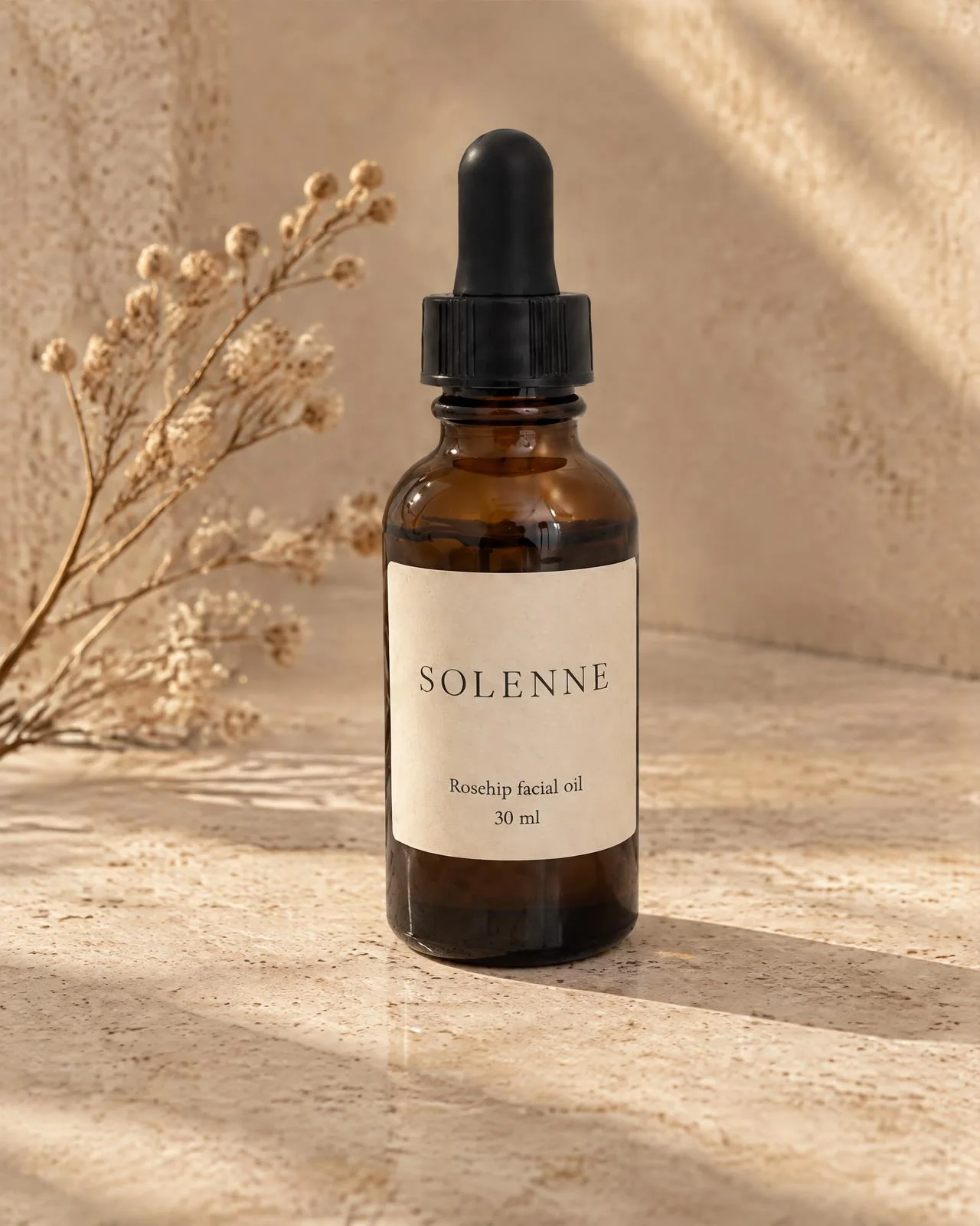 The rosehip oil bottle on travertine with long window shadows and a dried flower sprig.